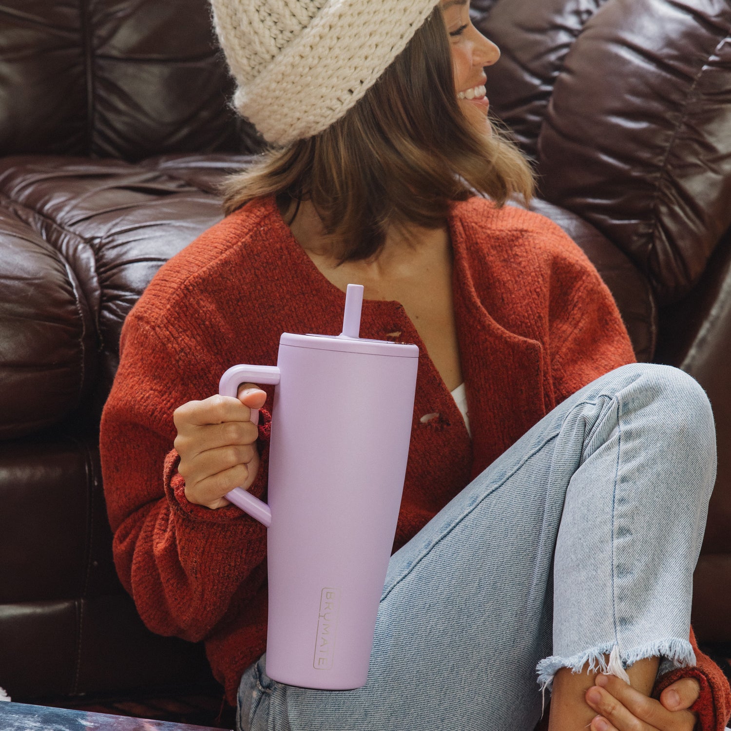 Woman sitting on a couch holding a light purple Era 40oz tumbler, wearing a red sweater and beige beanie.