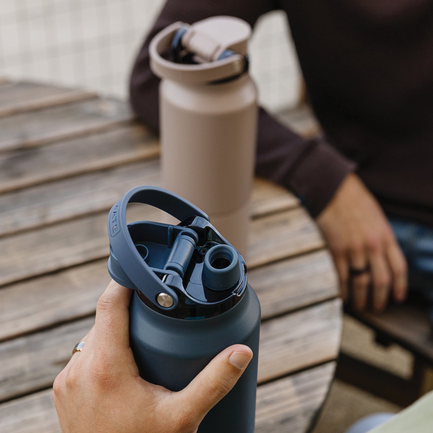 Person holding a blue Rise 35oz water bottle  with a beige Rise 25oz water bottle in the background on a wooden table.