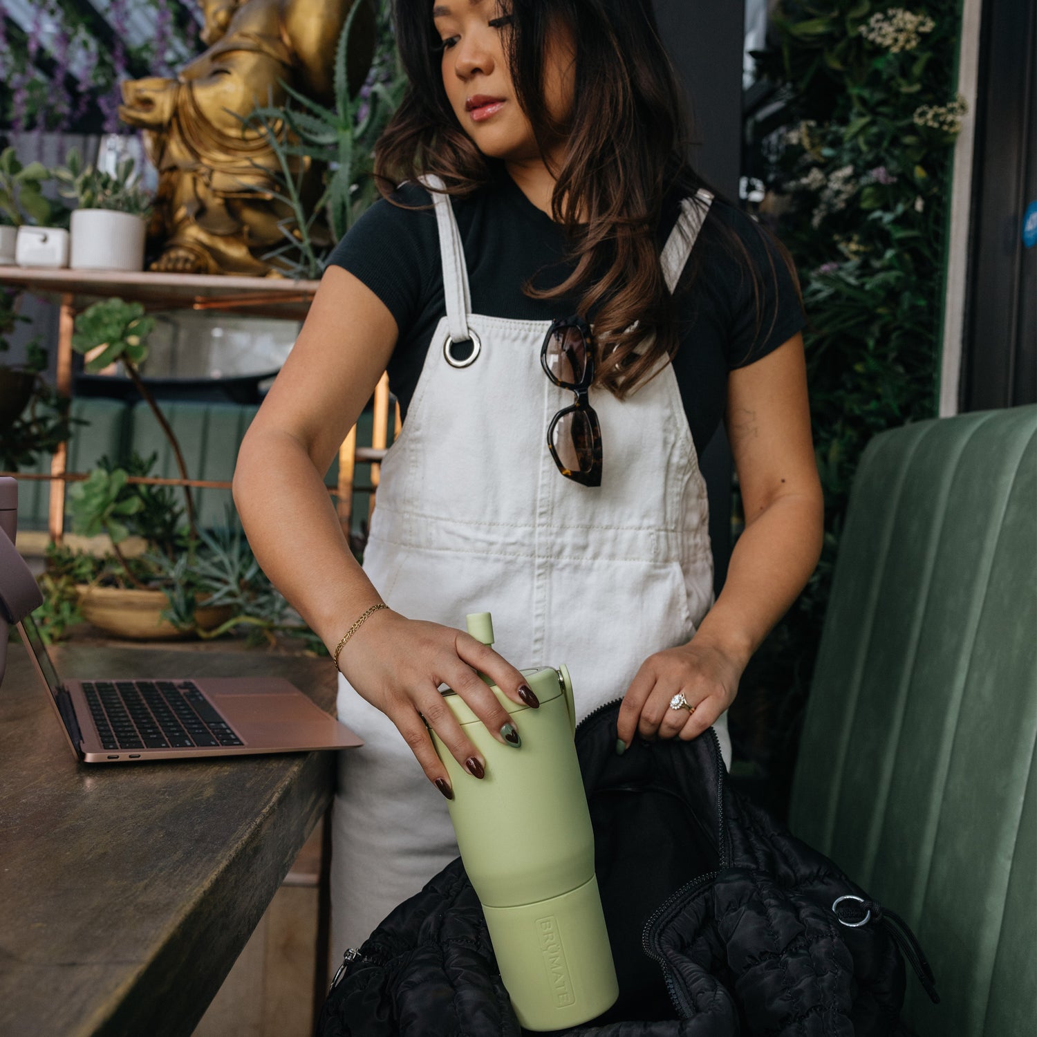 Woman in white overalls holding a green Resa 25oz tumbler in a casual setting with plants and a laptop.
