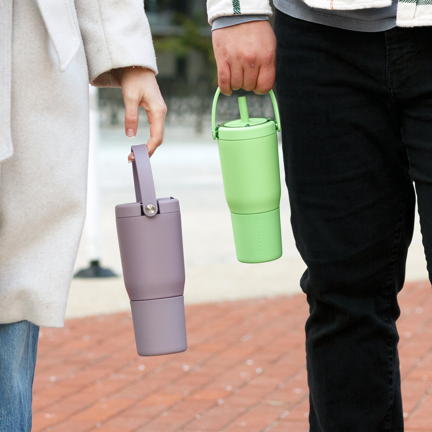 Two people holding a purple and a green Resa tumblers on a brick pavement.