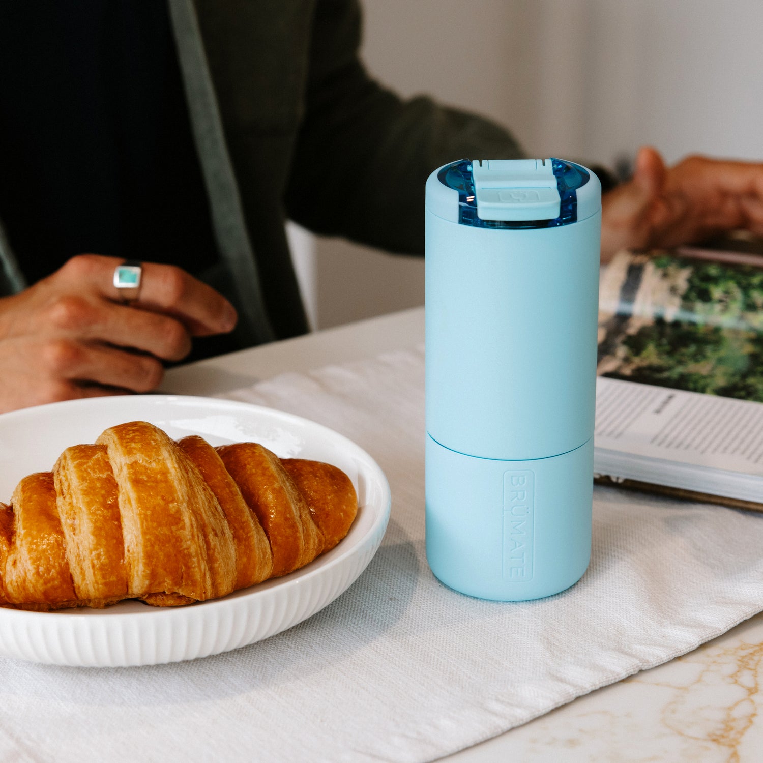 Light blue Strova 18oz coffee tumbler on a table with croissants and a book