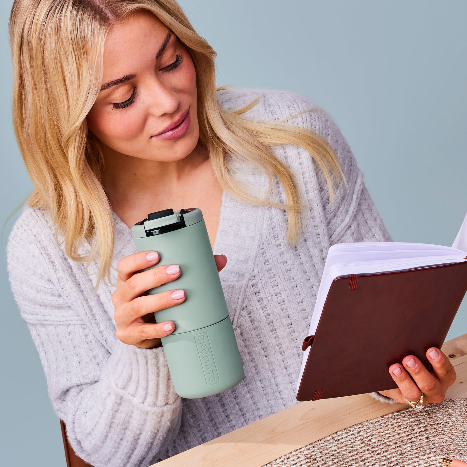 Woman holding a green Strova 18oz coffee tumbler and a book against a light blue background