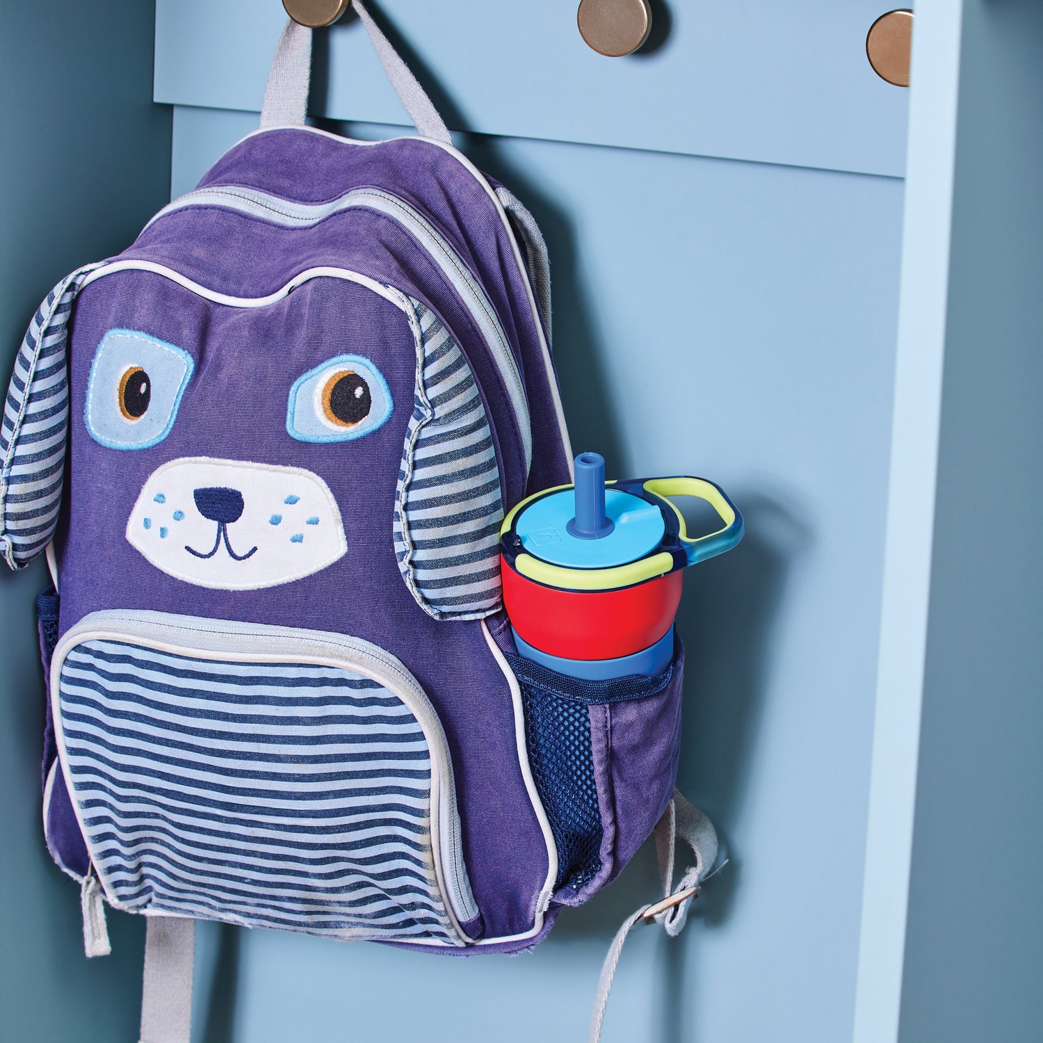 Children's backpack with dog face design and blue and red Leap 12oz tumbler attached to a blue cabinet.